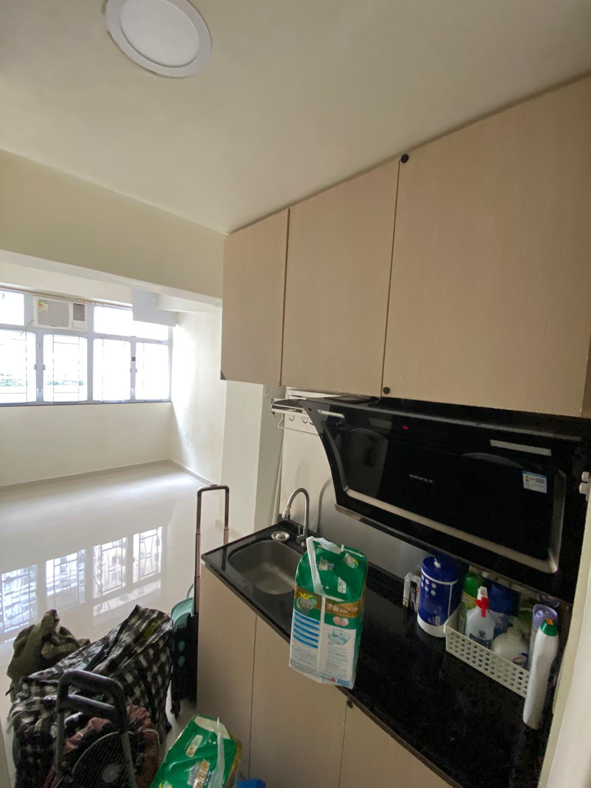 a well-lit, vacant studio apartment with a kitchen and some bags and adult diapers in the foreground, an empty living space, and a row of windows in the back