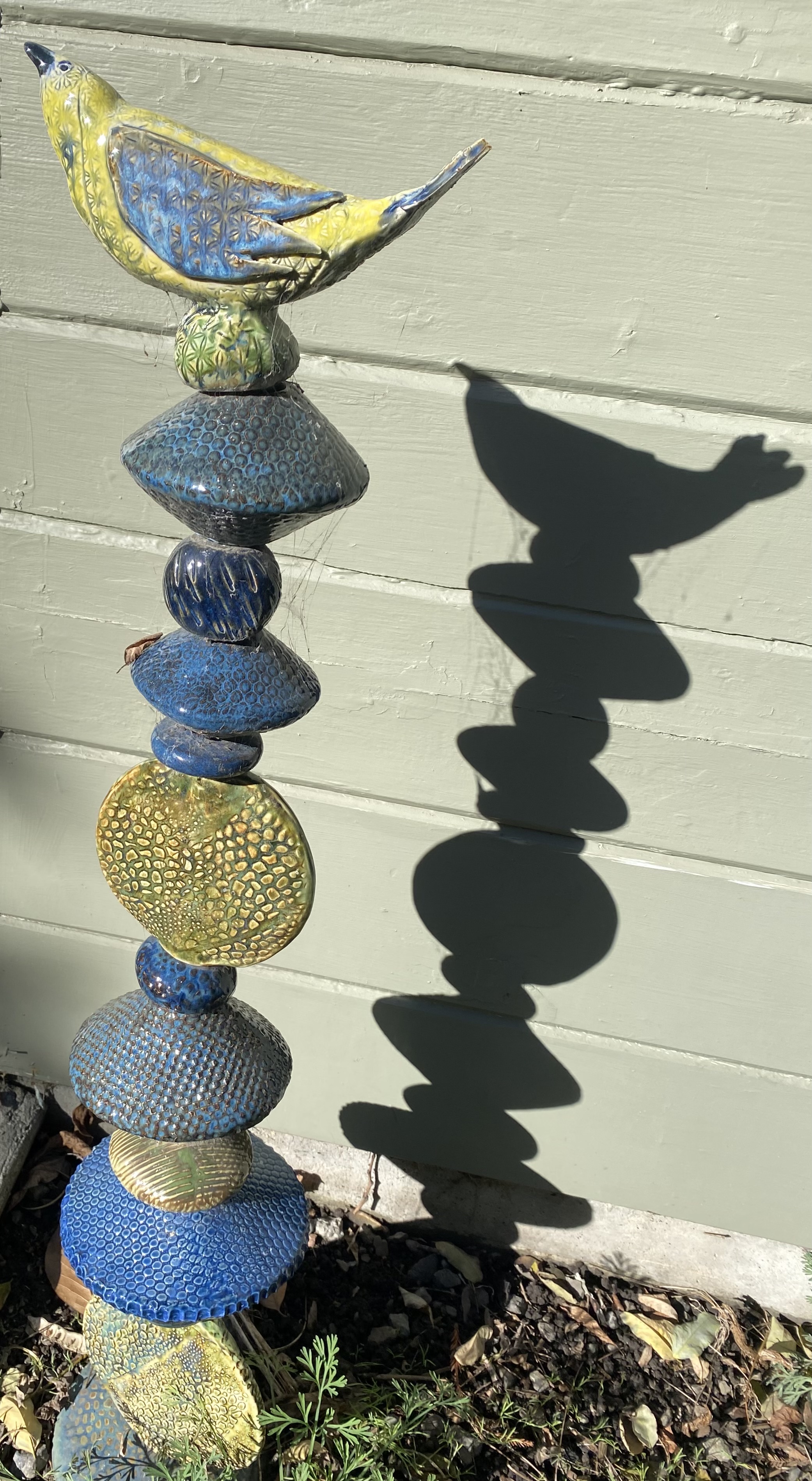 A ceramic sculpture of a bird sitting on top of a column of irregularly shaped discs, bicones and spheres.  Yellow and blue with touches of green.