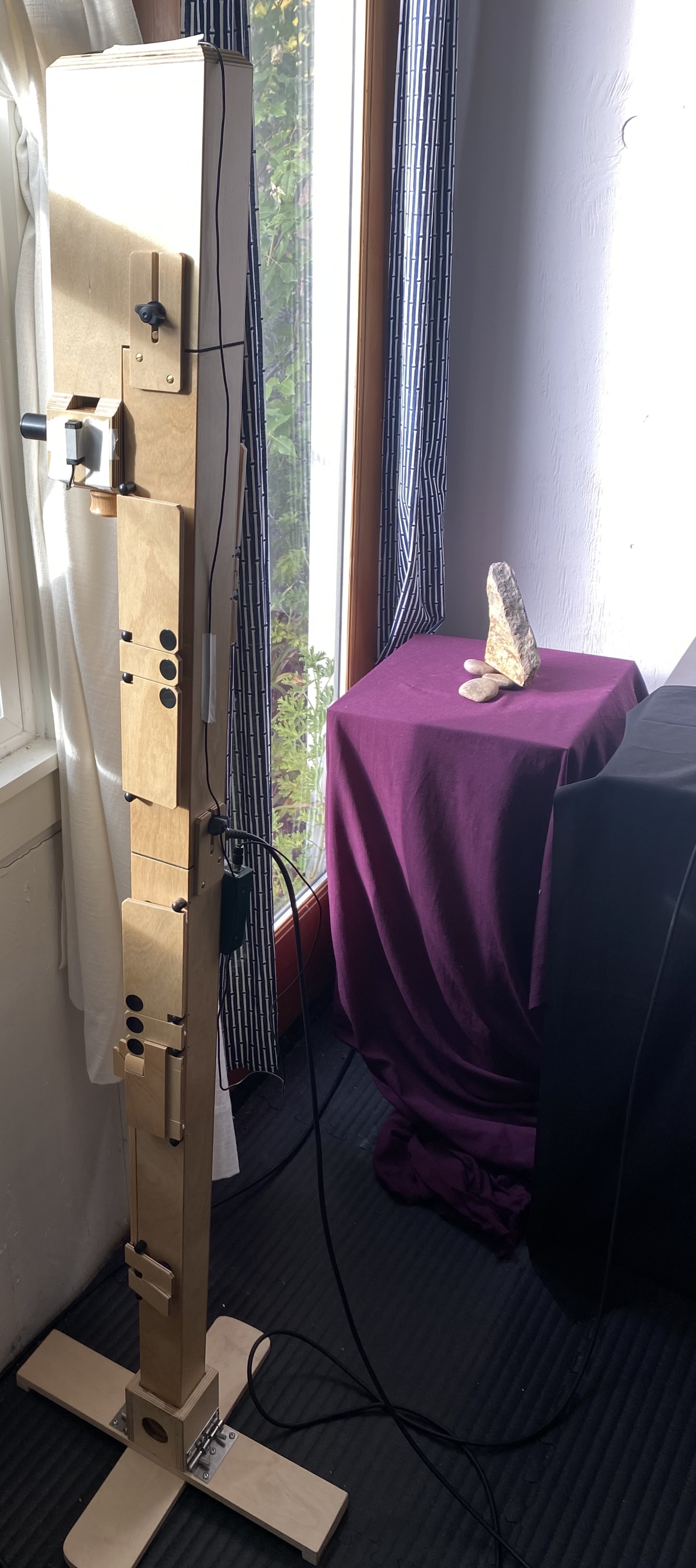 A contrabass Paetzold recorder by the window, rectangular wooden blocks with round black pads and a black mouthpiece.  A triagular rock with two smaller rocks on a table in the back with a burgundy table cloth.