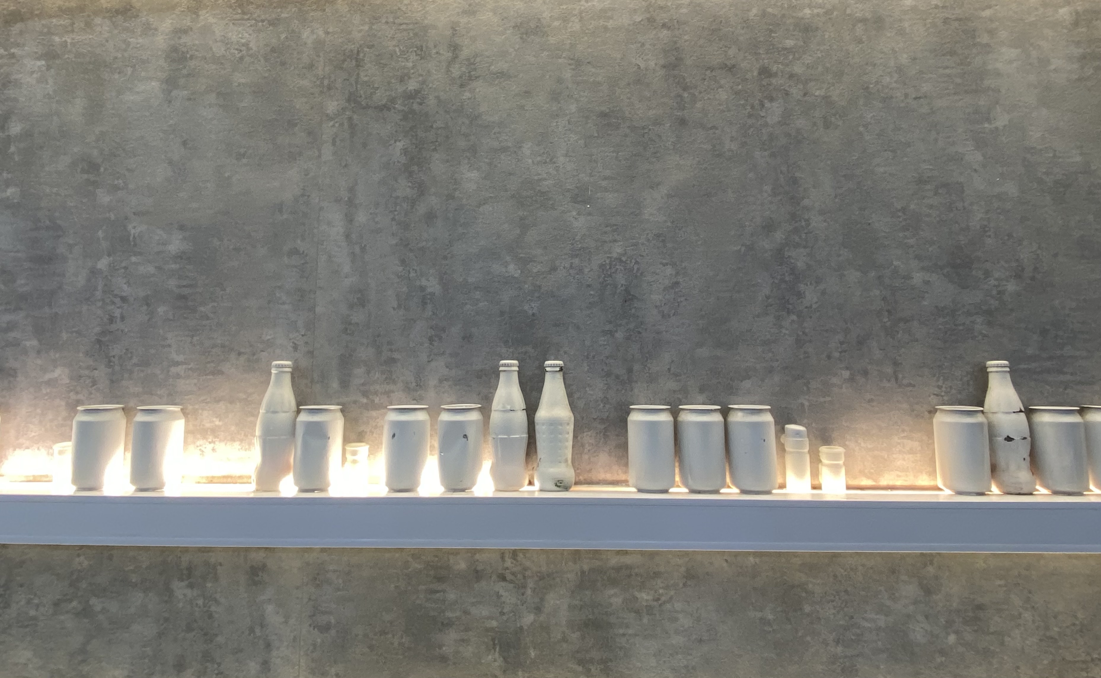 a row of soda can and bottles, salt and pepper shakers painted white, lined up on a shelf, lighted from below