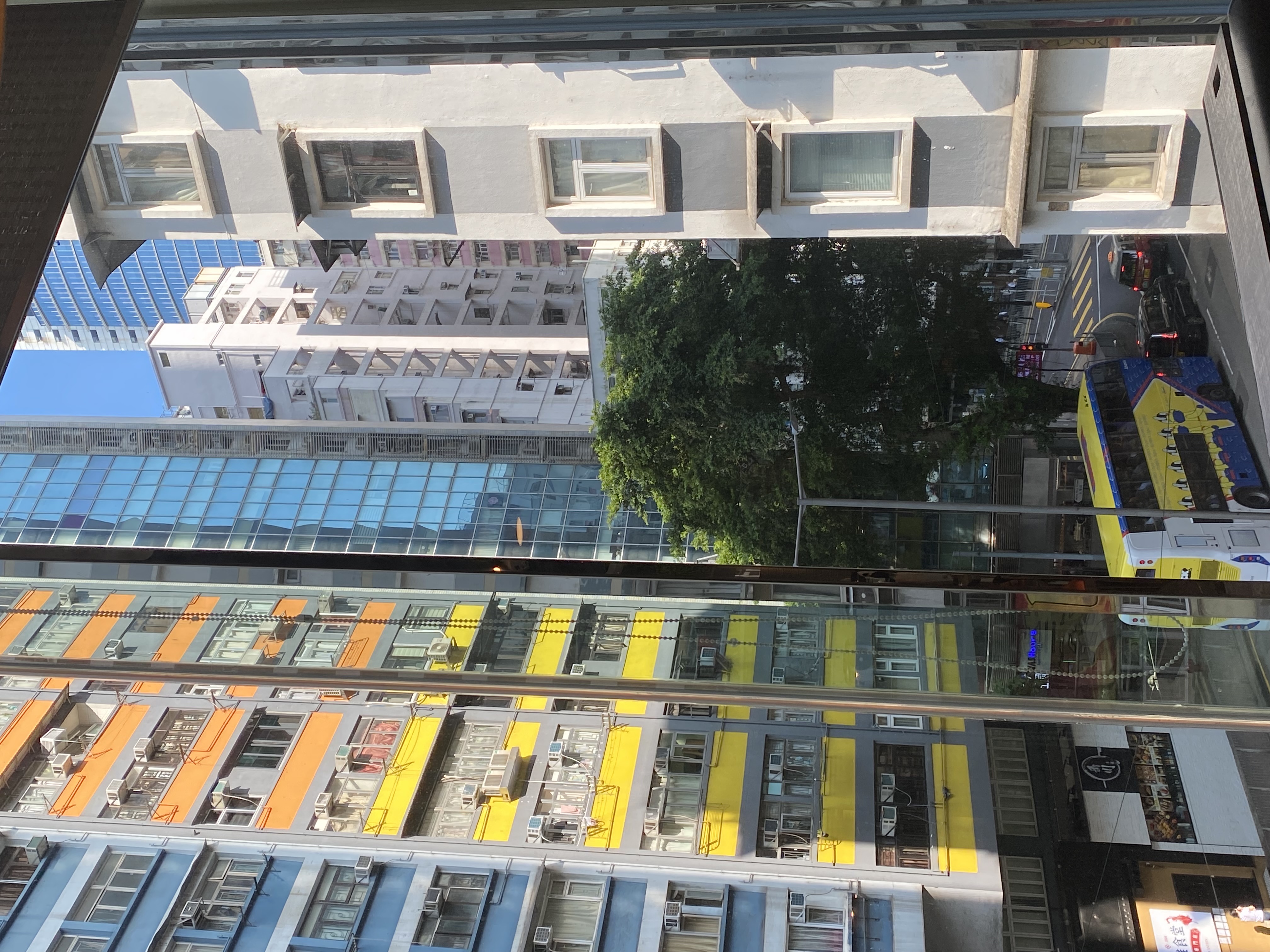 from a window, a tree across the street behind a double decker bus and taxis, next to a building with yellow, orange and blue panels, surrounded by many other tall buildings